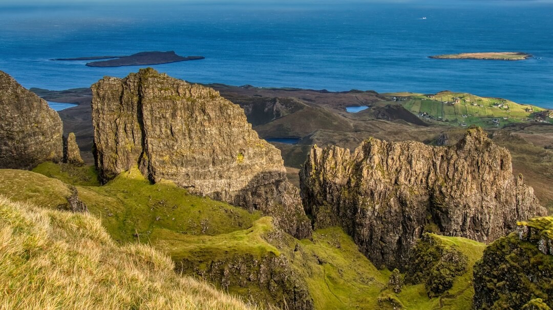A green plateau surrounded by jagged rock formations and a blue sea in the background