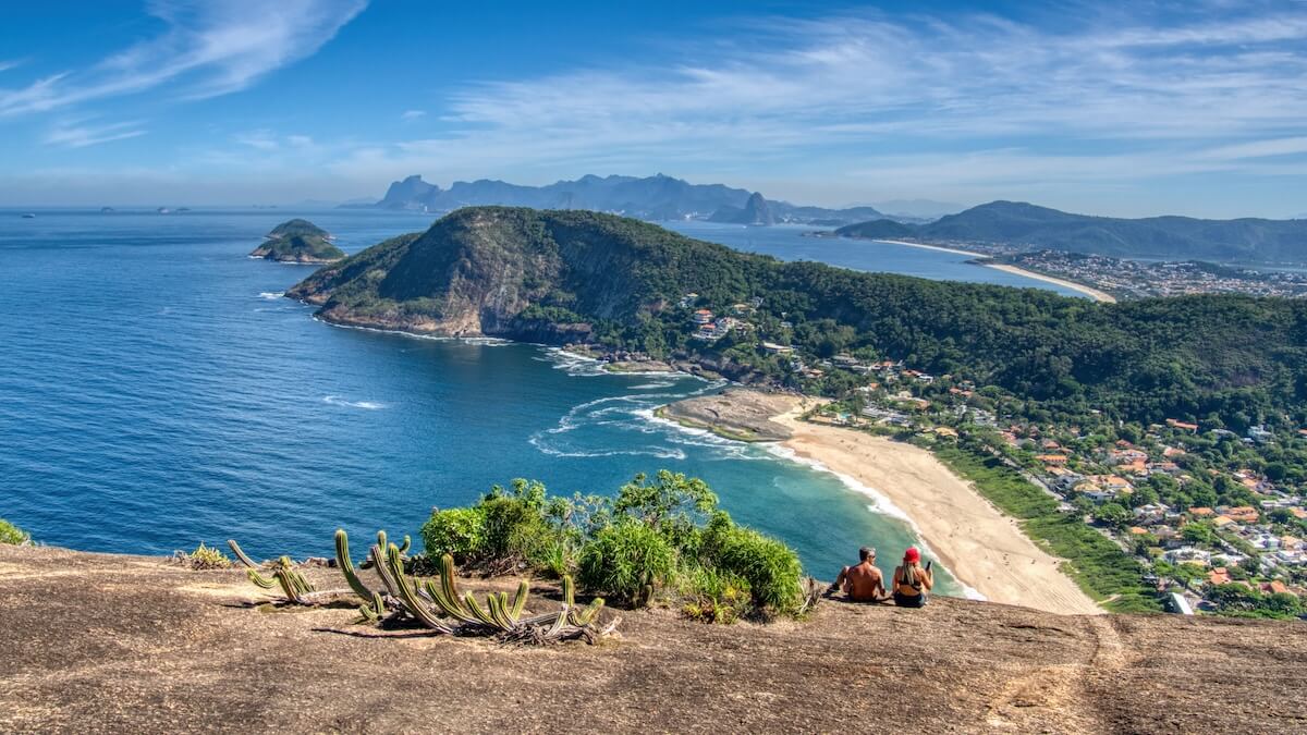 Two people sit on the edge of a cliff overlooking a beach and mountains behind